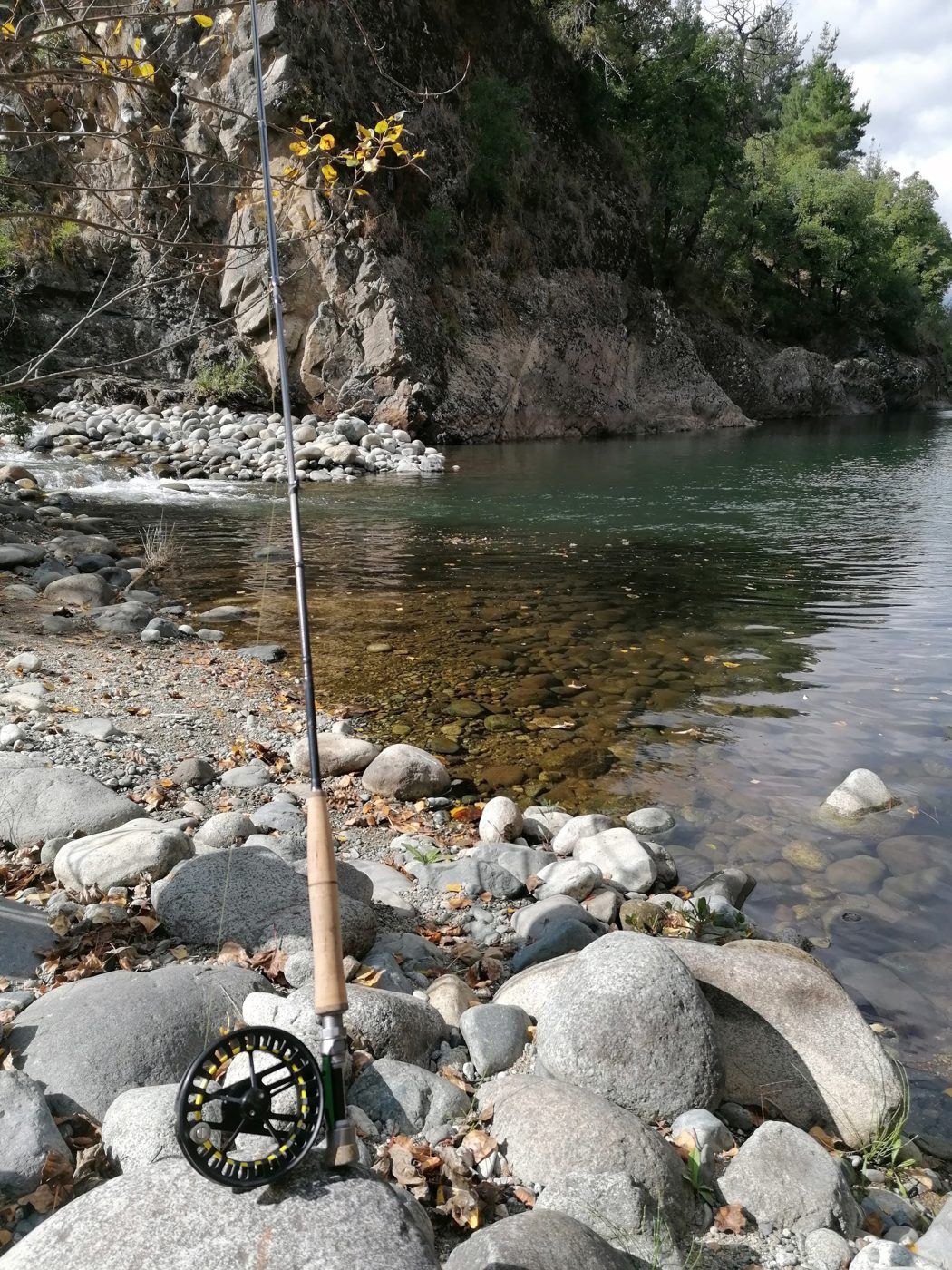 Vara de pesca com mosca e carretilha apoiadas nas pedras à beira de um rio de águas cristalinas