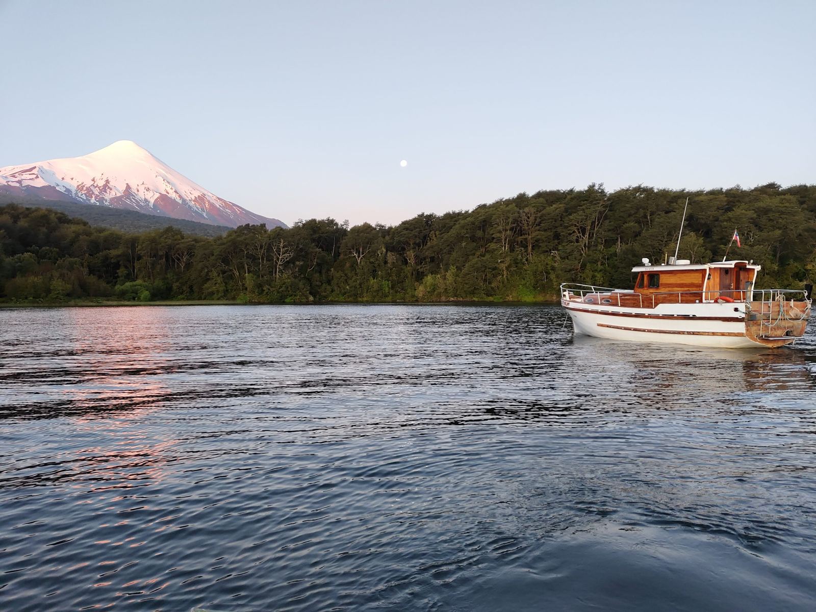 Iate Lithium ancorado ao entardecer, com o vulcão Osorno nevado e a lua cheia sobre a floresta